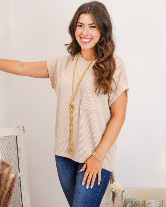 Woman wearing a beige textured short sleeve top with front pocket, styled with a long gold necklace and dark denim jeans from MaKenzie Layne Boutique.