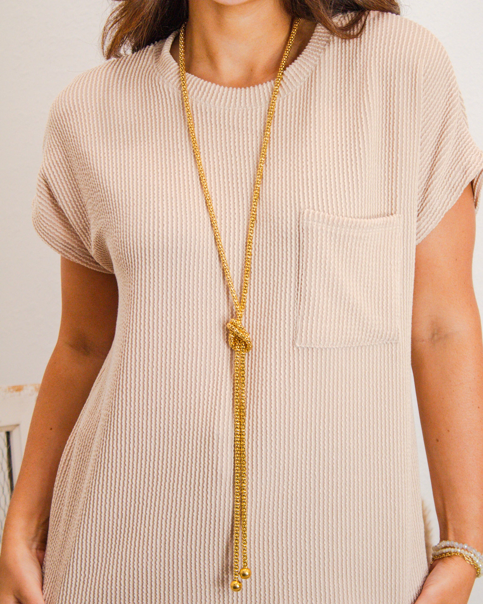 Close-up of beige waffle-knit short sleeve top with chest pocket, styled with a long knotted gold chain necklace from MaKenzie Layne Boutique.