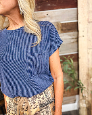 Women's navy blue ribbed knit short sleeve top with a relaxed fit, round neckline, and front chest pocket, styled with brown paisley print pants at MaKenzie Layne Boutique.