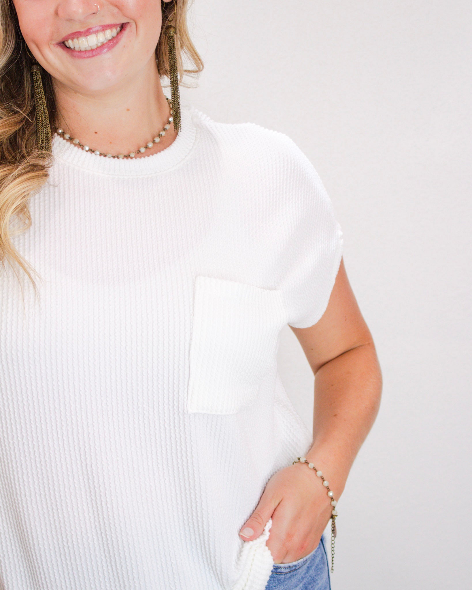 Close-up of woman wearing a textured white ribbed short sleeve top with a front pocket, paired with light wash denim jeans and beaded accessories.