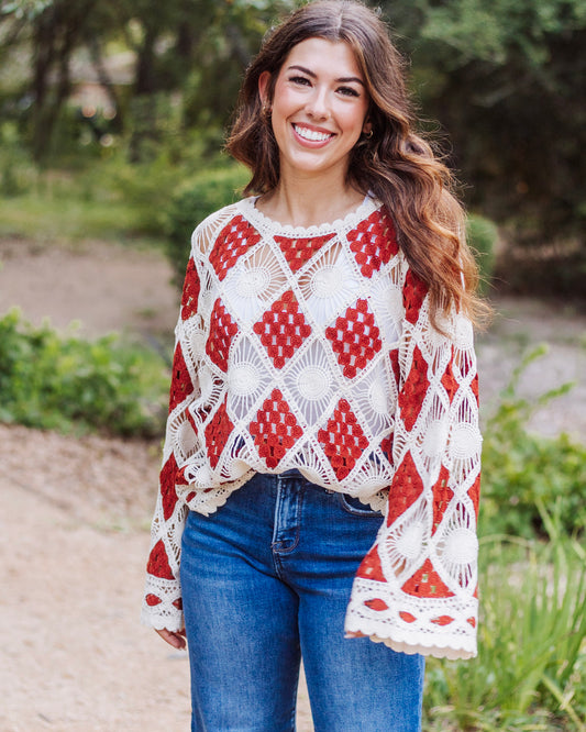 Woman wearing a long-sleeve crochet sweater with red and cream diamond patterns, paired with blue jeans, smiling outdoors in a natural setting.