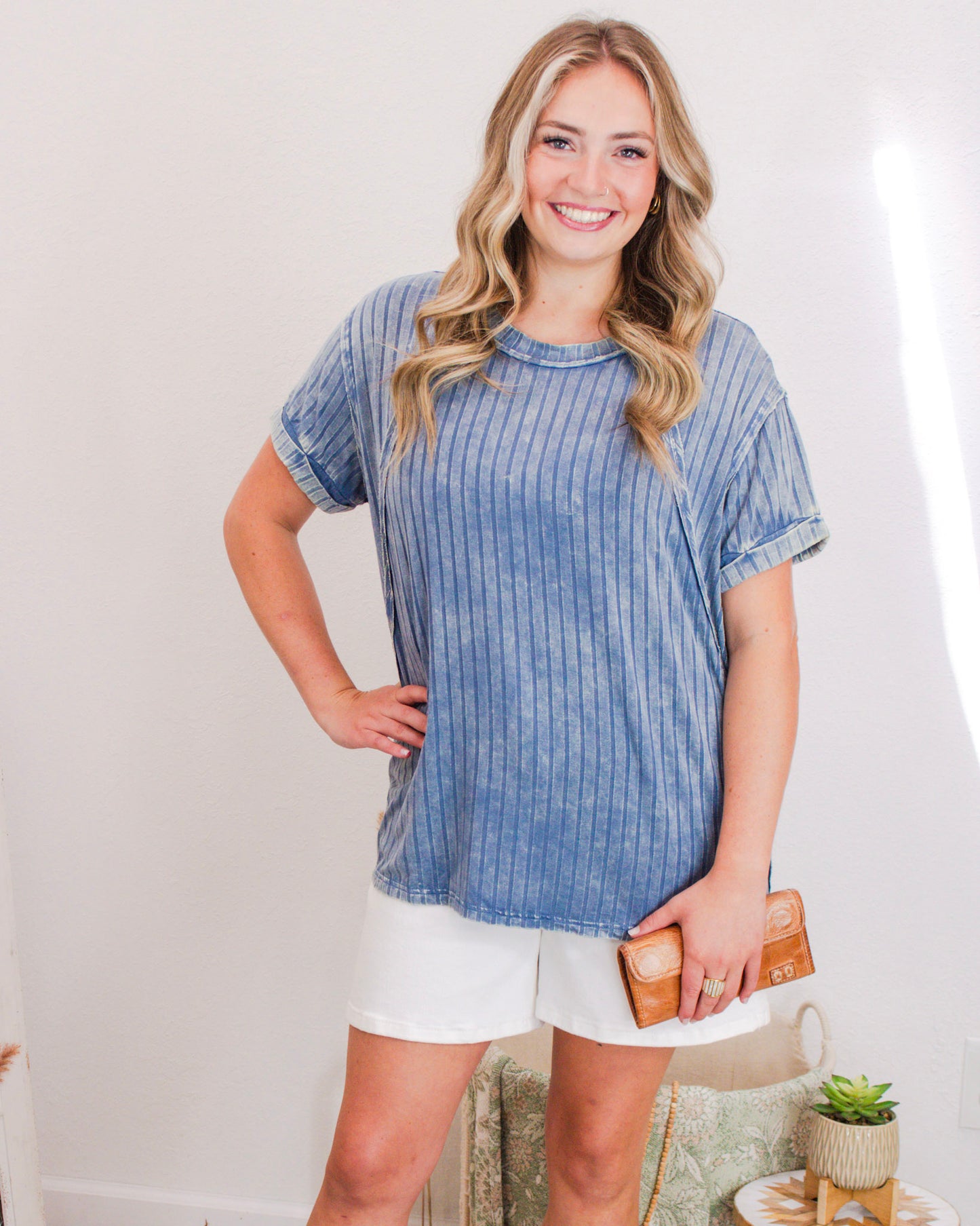 Smiling woman wearing a blue pinstripe short-sleeve top with white shorts and holding a tan wallet.