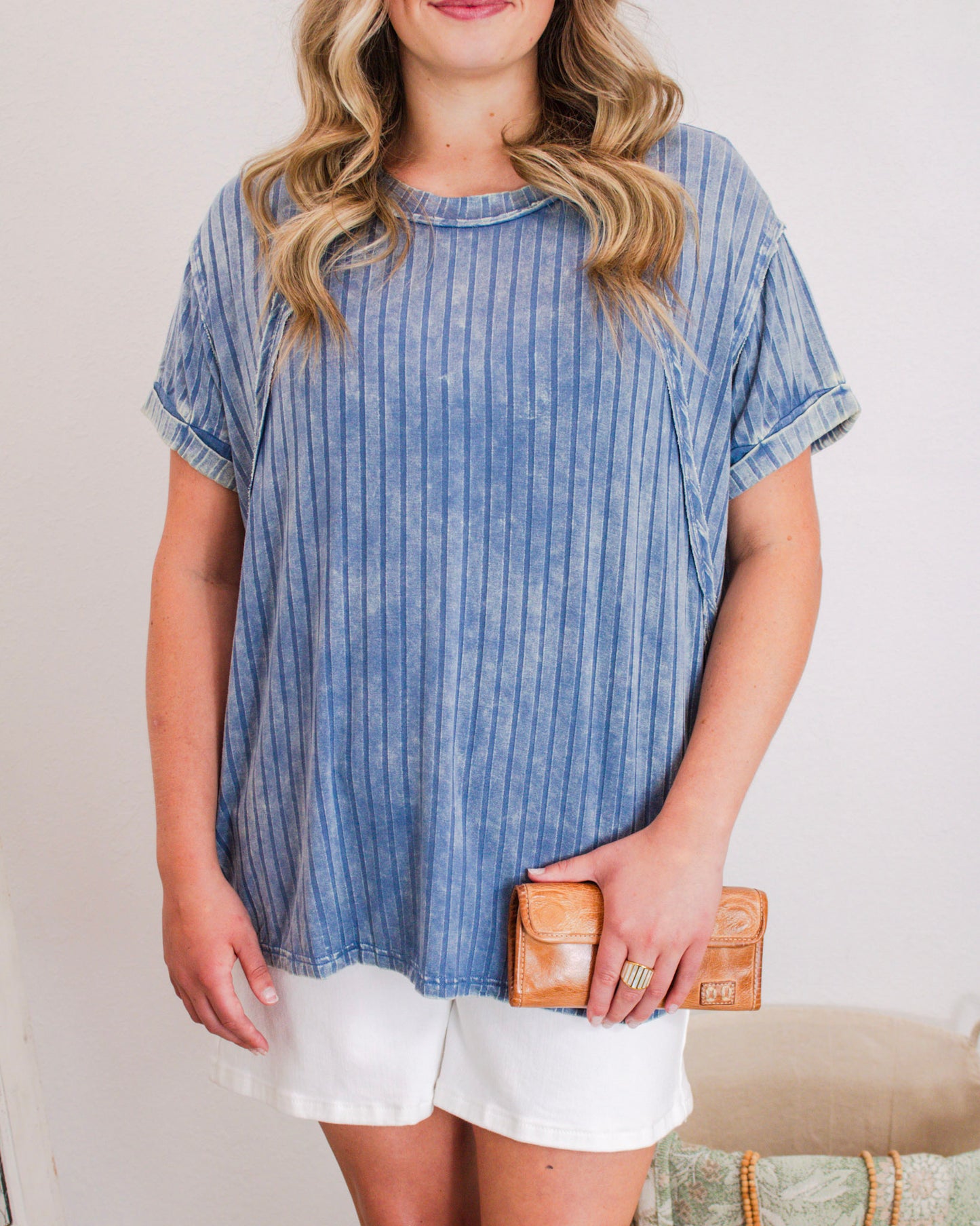Woman wearing a relaxed blue pinstripe top with cuffed sleeves and white shorts, holding a tan leather wallet.