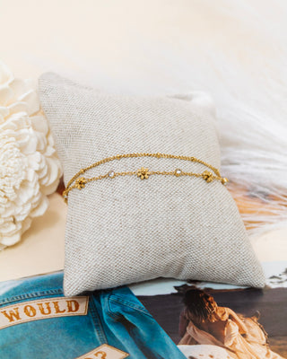 Set of delicate gold chain bracelets featuring small bead and floral accents, artfully displayed on a neutral fabric cushion with decor accents in background