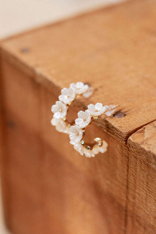 Pair of gold hoop earrings adorned with delicate white flower accents and pearl centers, displayed on a rustic wooden surface. Feminine and elegant floral jewelry for spring and summer styling.
