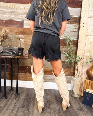 Back view of women's black lace-trim shorts with elastic waistband, styled with a charcoal graphic tee and cream western cowboy boots at MaKenzie Layne Boutique.