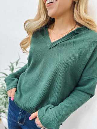 Woman wearing a forest green V-neck sweater with layered gold necklaces and dark blue denim jeans, standing against a white textured wall