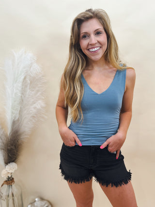 Woman wearing a fitted blue V-neck tank top with black distressed fringe hem shorts, styled for a casual summer look against a neutral backdrop.