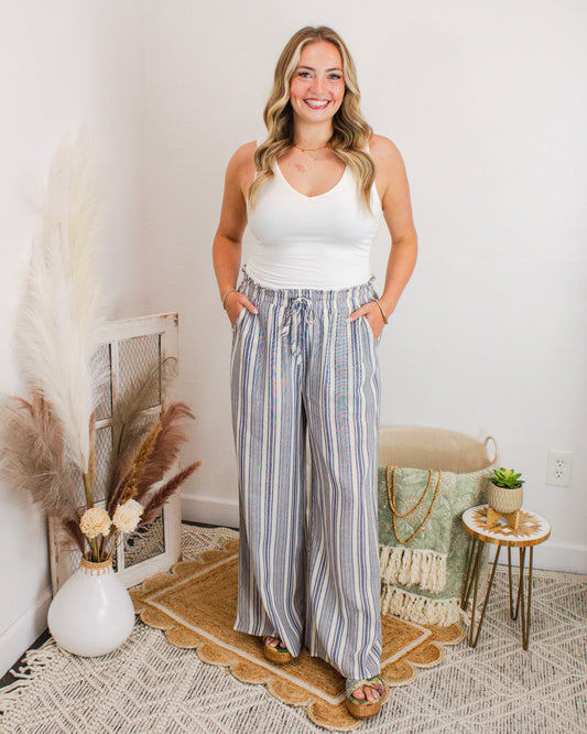 Woman wearing flowy blue and white striped wide-leg pants with a drawstring waist, styled with a fitted white tank top and sandals for a relaxed summer outfit.