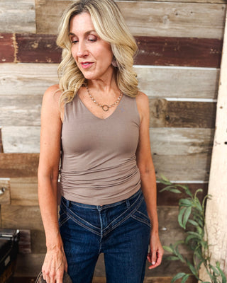 Women's taupe sleeveless V-neck tank top with flattering ruched sides, styled with dark wash high-waisted western seam jeans and a delicate gold necklace for a casual chic outfit.
