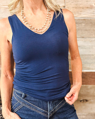 Close-up of women's navy blue sleeveless V-neck tank top featuring a fitted silhouette and smooth stretch fabric, styled with dark wash high-waisted western jeans and a layered beaded necklace.
