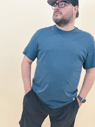 Man wearing a blue performance t-shirt and black athletic shorts with hands in pockets, styled with a watch and hat for a relaxed, casual summer look.