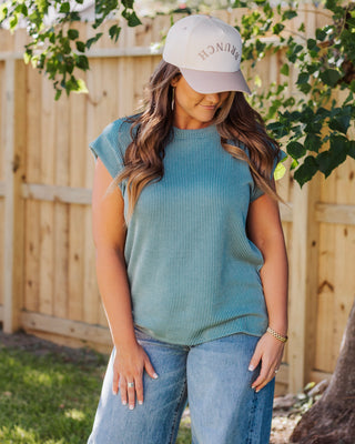 Woman wearing a blue ribbed knit sleeveless top paired with high-waisted blue jeans and a beige baseball cap, styled for a casual outdoor look.