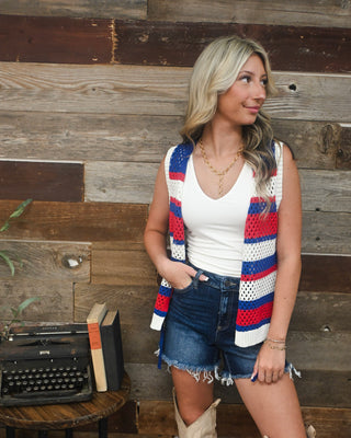 Women’s patriotic crochet knit vest in red, white and blue layered over a cream tank with denim shorts for trendy Americana summer boutique style.