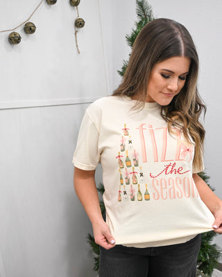 Woman wearing an ivory holiday graphic T-shirt with champagne bottle Christmas tree design and the phrase ‘Fizz the Season,’ styled with dark jeans.