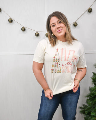 Woman wearing an ivory graphic T-shirt with champagne bottle Christmas tree design and the phrase ‘Fizz the Season,’ styled with dark-wash jeans.