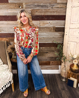 Women’s cream floral print blouse with vibrant red, pink and orange flowers, ruffle shoulders and long sleeves, styled with high-rise wide leg jeans for fall.