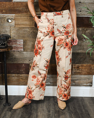 Women’s cream floral wide-leg pants with a high-rise fit and rust-toned botanical print, styled with a brown top and neutral studded mules.