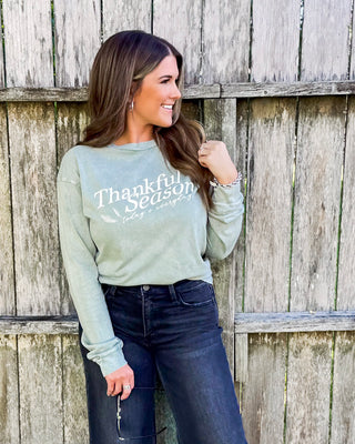 Woman wearing a sage green “Thankful Season” sweatshirt paired with dark jeans, styled for fall with a casual, cozy boutique look against a rustic wooden backdrop.