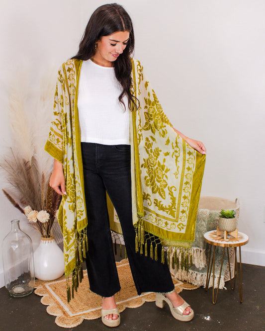 Woman modeling an olive green and ivory floral kimono with tassel fringe, styled over a white sleeveless top and black wide-leg jeans in a boho-style setting.