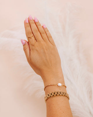 Hand with pink nails wearing a gold open heart ring, a dainty pearl bead bracelet, and a chunky gold watch link bracelet against a soft blush feathered background.