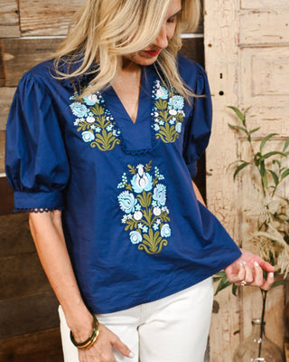 Royal blue embroidered blouse with light blue and olive floral detailing, V-neckline and puff sleeves, styled with white jeans for an elevated boutique look.