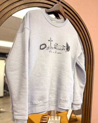 Women’s light gray crewneck sweatshirt featuring “He is Risen” graphic with cross, lamb, crown of thorns, and palm branch for a faith-inspired boutique look