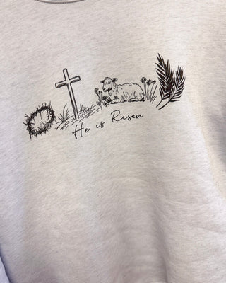 Women’s cream graphic tee featuring “He is Risen” with cross, lamb, crown of thorns, and palm branch illustration for a faith-inspired boutique look