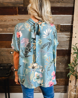 Women’s sage blue floral blouse featuring a relaxed fit, botanical print and feminine tie-back neckline, styled with denim for an effortless boho boutique look.