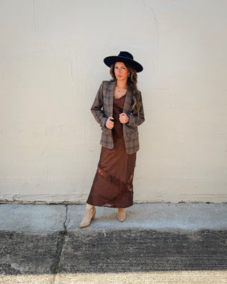 Women's fall western chic outfit featuring a brown and taupe plaid blazer layered over a chocolate brown lace-trim satin midi dress, styled with a black wide-brim cowboy hat and beige western ankle boots.