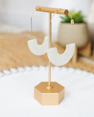 Pair of modern U-shaped white acrylic earrings with gold wire hooks displayed on a gold earring stand, styled against a neutral boho background.