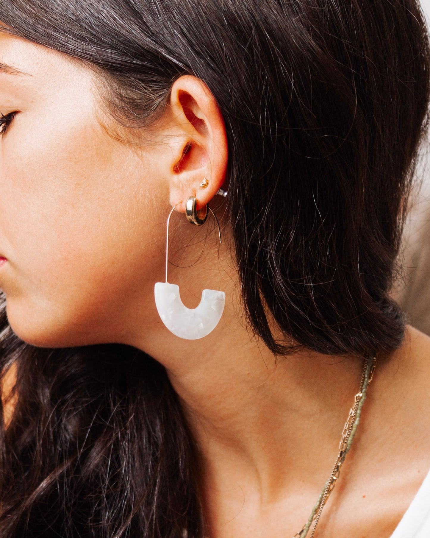 Close-up of a woman wearing modern gold wire earrings with a white translucent U-shaped acrylic charm, styled with layered gold necklaces and natural makeup.