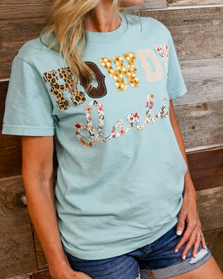 Sage green “Howdy Darlin” patchwork graphic tee with floral and western prints, styled with distressed denim shorts, casual women’s boutique outfit against rustic wood backdrop