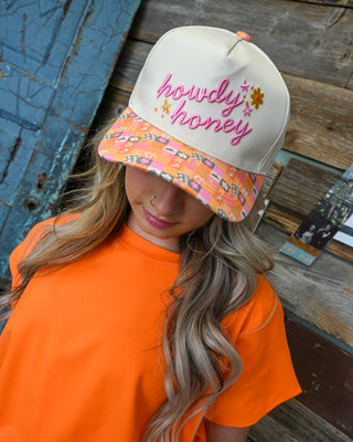 Women’s cream “Howdy Honey” trucker hat with pink embroidered lettering and multicolor patterned brim, styled with a bright orange top for a playful western boutique look