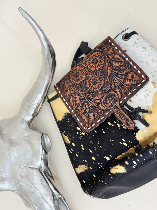 Tooled leather and cowhide backpack with floral design flap and gold splatter accents styled beside silver bull skull for a bold western-inspired fashion look.