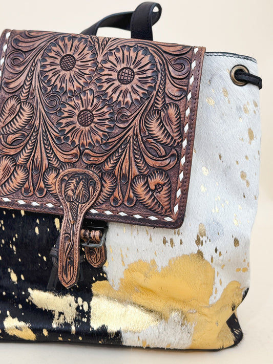 Western cowhide backpack with tooled leather floral flap and gold splatter detailing—boho-chic and rustic accessory perfect for everyday or festival style.