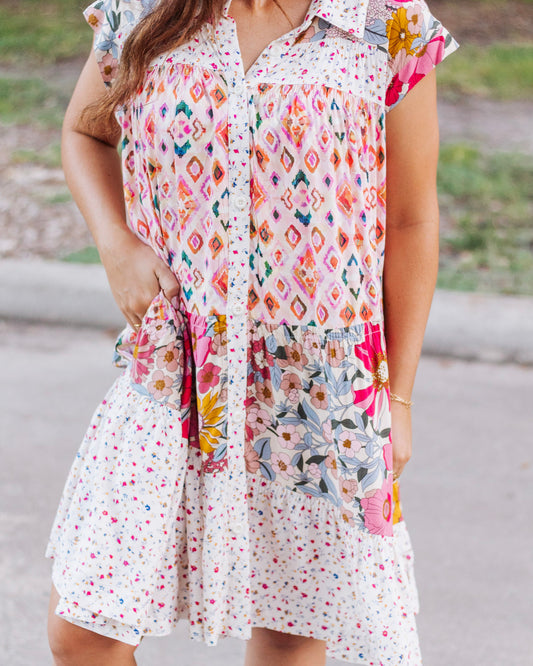Close-up of a boho patchwork mini dress with floral, geometric, and polka dot patterns in pink, orange, and cream tones, featuring button-down front and side pockets.