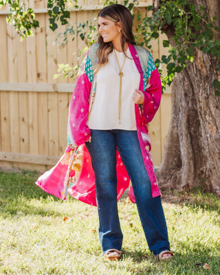 Woman wearing a vibrant pink and teal patterned kimono layered over a white top with dark blue jeans and sandals, styled for a relaxed boho-chic outfit.