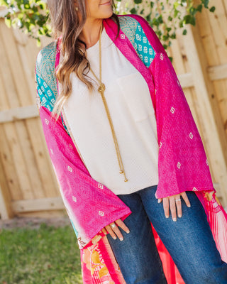 Woman wearing a bright pink and teal patterned kimono over a white textured top with blue jeans, styled with a long gold necklace for a boho chic look.