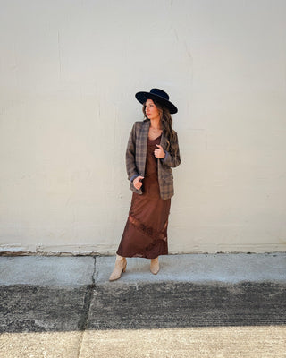 Women's western-inspired fall outfit featuring a brown and taupe plaid blazer over a chocolate brown satin midi dress with lace detailing, styled with a black wide-brim cowboy hat and beige pointed-toe ankle boots.