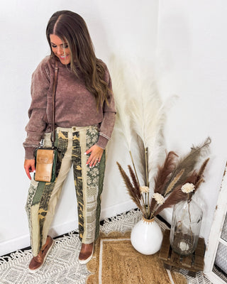 Woman wearing patchwork print high-rise pants in olive and cream tones, styled with a washed brown sweatshirt, leather crossbody, and slip-on sneakers.