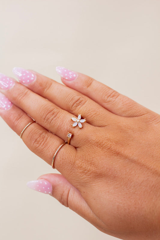 Close-up of hand wearing a gold open ring featuring a white floral design on one end and a small crystal accent on the other. Dainty and feminine adjustable ring perfect for spring and everyday wear.