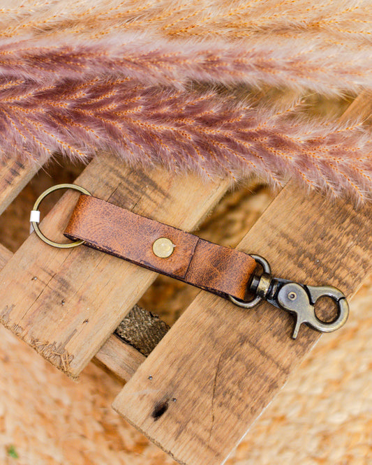 Handcrafted brown leather keychain with brass hardware and swivel clasp on rustic wooden surface – durable and stylish everyday accessory.