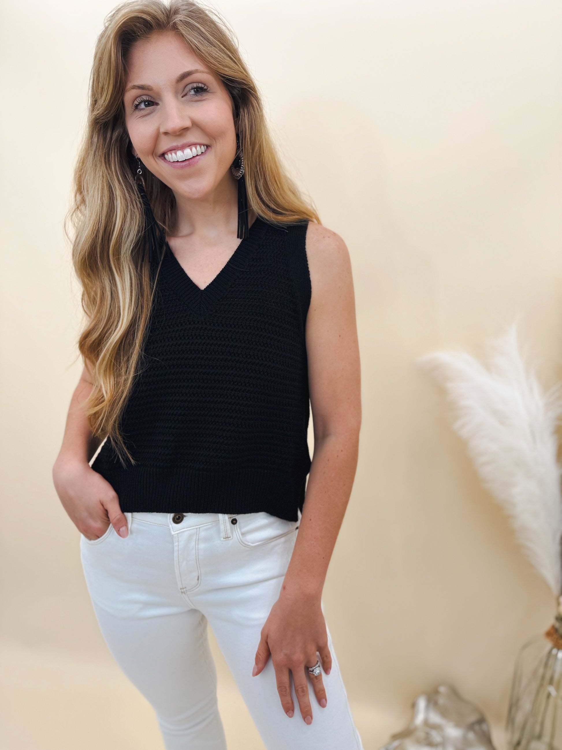 Woman wearing a sleeveless black knit sweater top paired with high-rise white skinny jeans, styled with long tassel earrings for a chic and modern look.