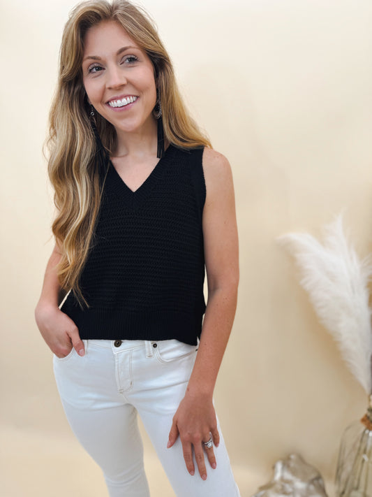 Woman wearing a sleeveless black knit sweater top paired with high-rise white skinny jeans, styled with long tassel earrings for a chic and modern look.