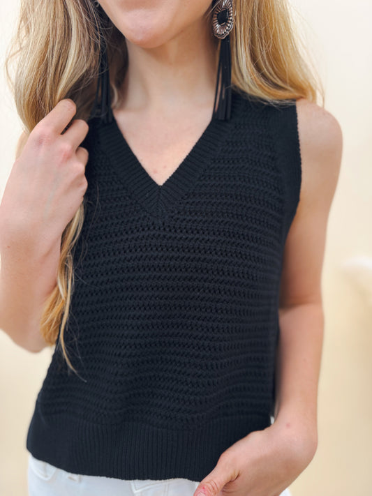 Close-up of a woman wearing a sleeveless black crochet knit sweater tank with a V-neckline, styled with long tassel earrings and white denim for a chic look.