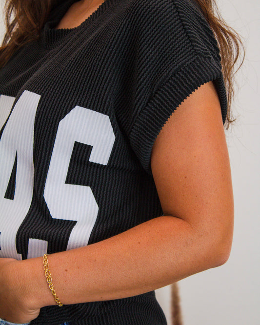 Close-up of a black ribbed knit "TEXAS" graphic top paired with a delicate gold chain bracelet on a woman's arm.