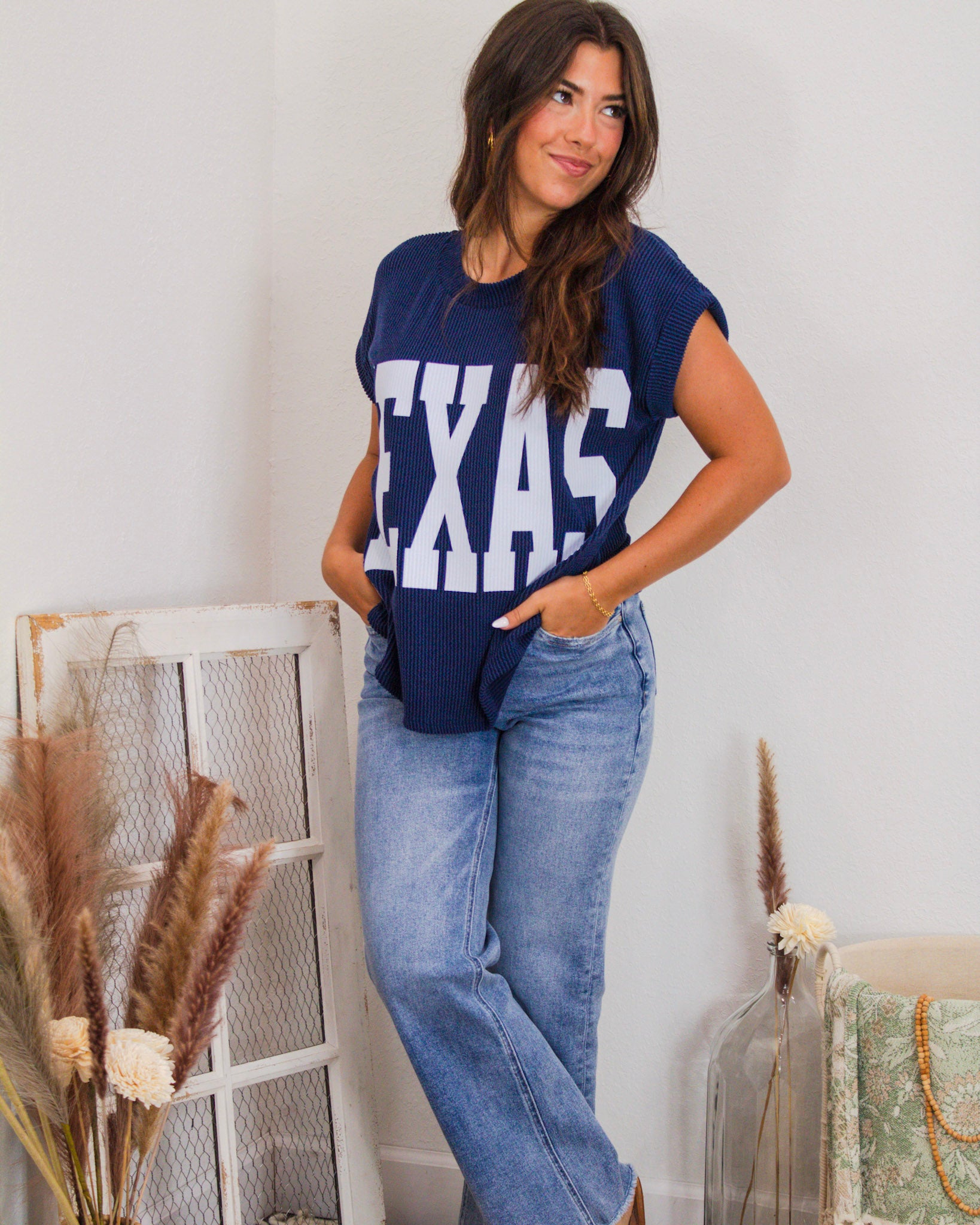 Woman wearing a navy ribbed knit graphic tee with bold white "TEXAS" lettering, styled with light-wash jeans and a gold chain bracelet.