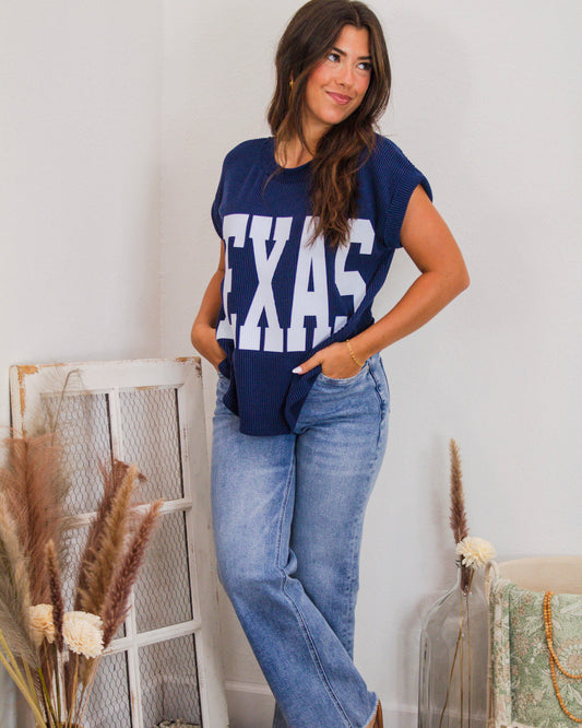 Woman wearing a navy ribbed knit graphic tee with bold white "TEXAS" lettering, styled with light-wash jeans and a gold chain bracelet.