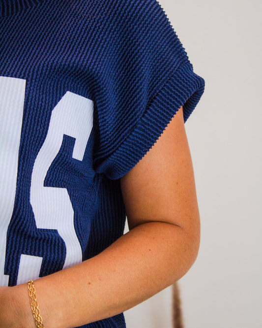 Close-up of a navy ribbed knit "TEXAS" graphic top paired with a delicate gold chain bracelet on a woman's arm.